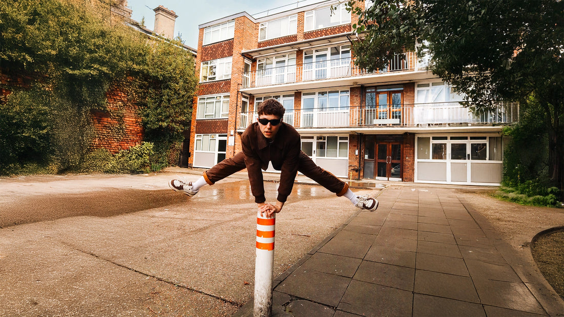 A man in a brown hoodie and sunglasses does a split leg pose on top of an orange-and-white striped bollard post in an outdoor courtyard, with a brick apartment building in the background.