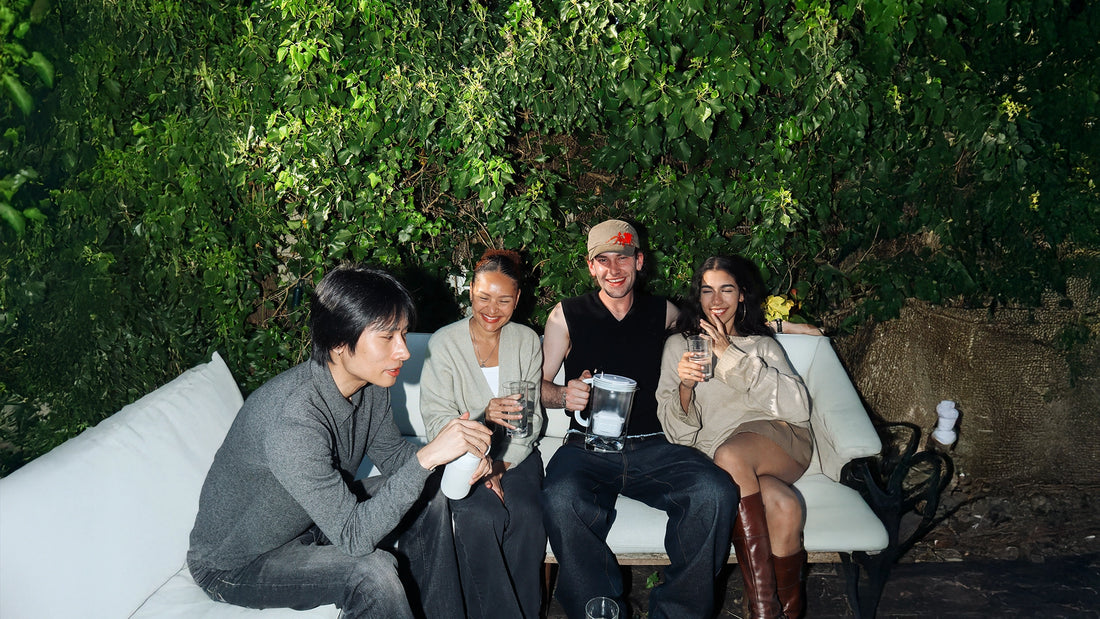 Group of four people sitting outdoors on a white sofa surrounded by green foliage, holding glasses of water, a BRITA water filter jug, and a BRITA water filter bottle during a social gathering.