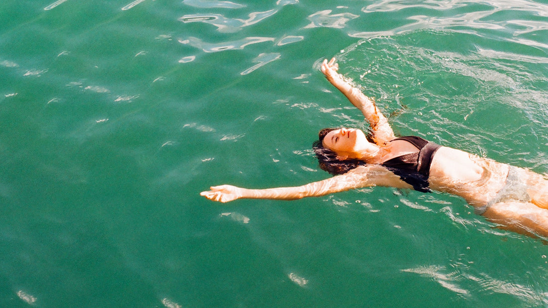 A smiling woman floating on her back in water, wearing a black bikini top and a light beige bikini bottom.