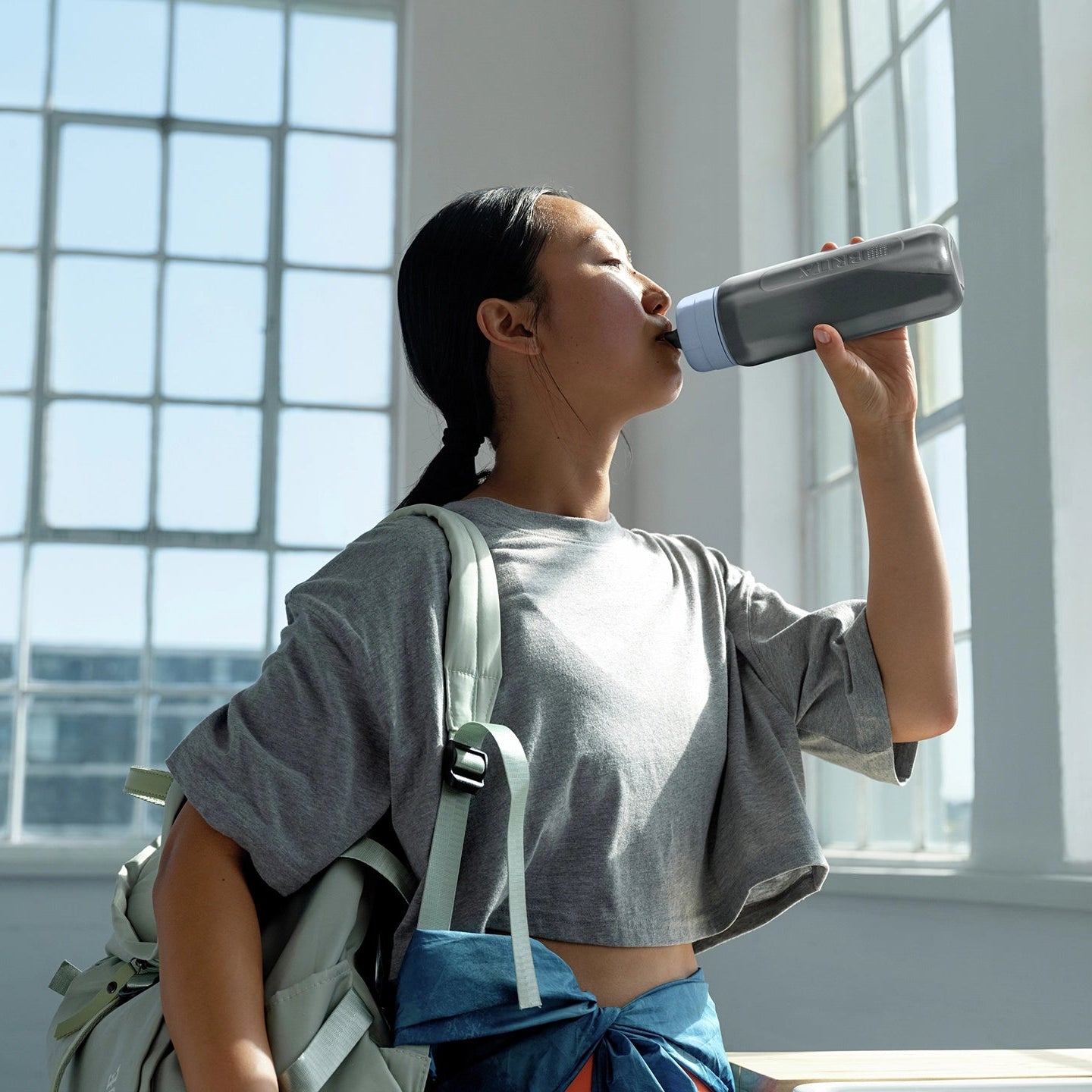 Woman drinking from a BRITA water filter bottle Model Active in a bright room with large windows.