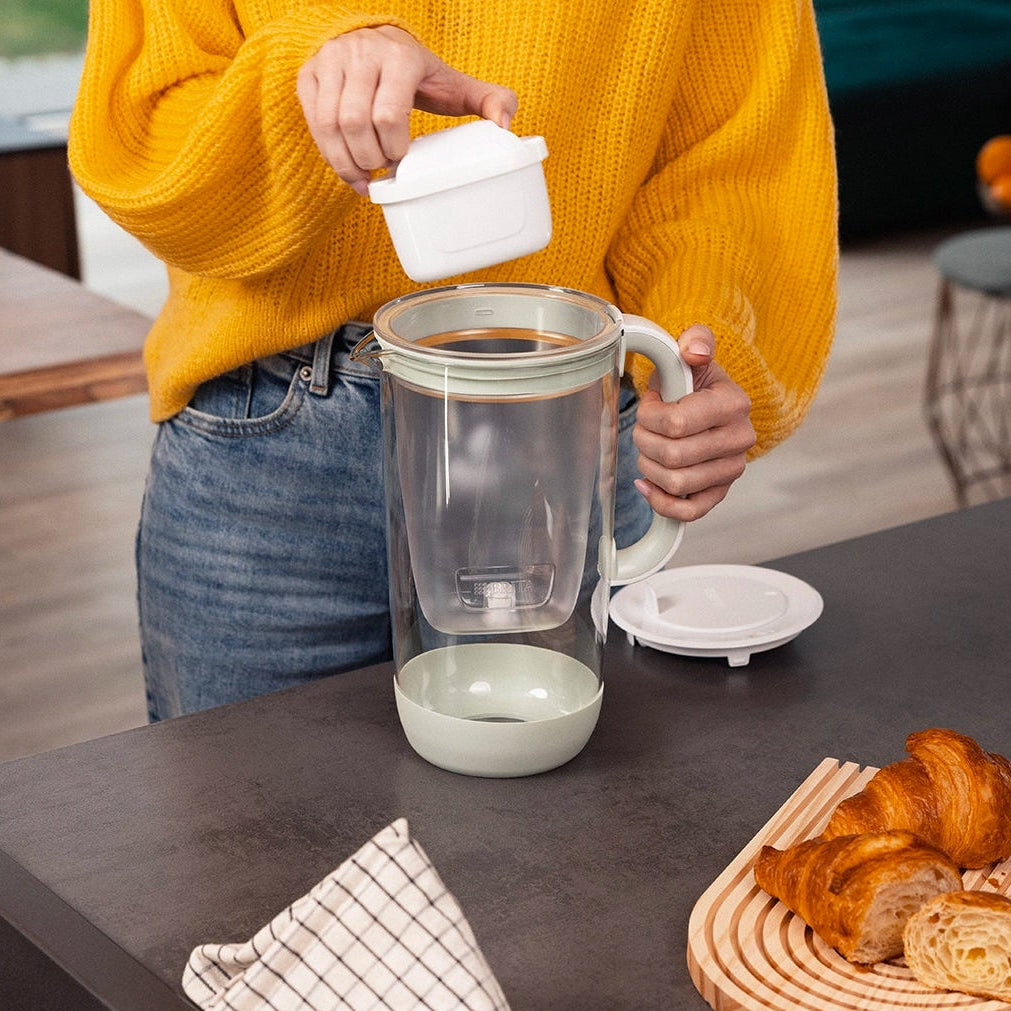 Person inserting a MAXTRA PRO filter cartridge into a green BRITA Glass Jug on a countertop.