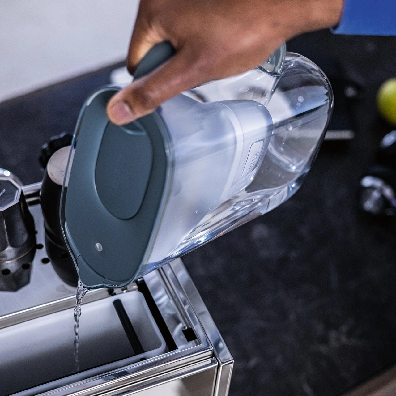 Person pouring filtered water from a blue BRITA Style Essential water filter jug into a coffee machine water tank.