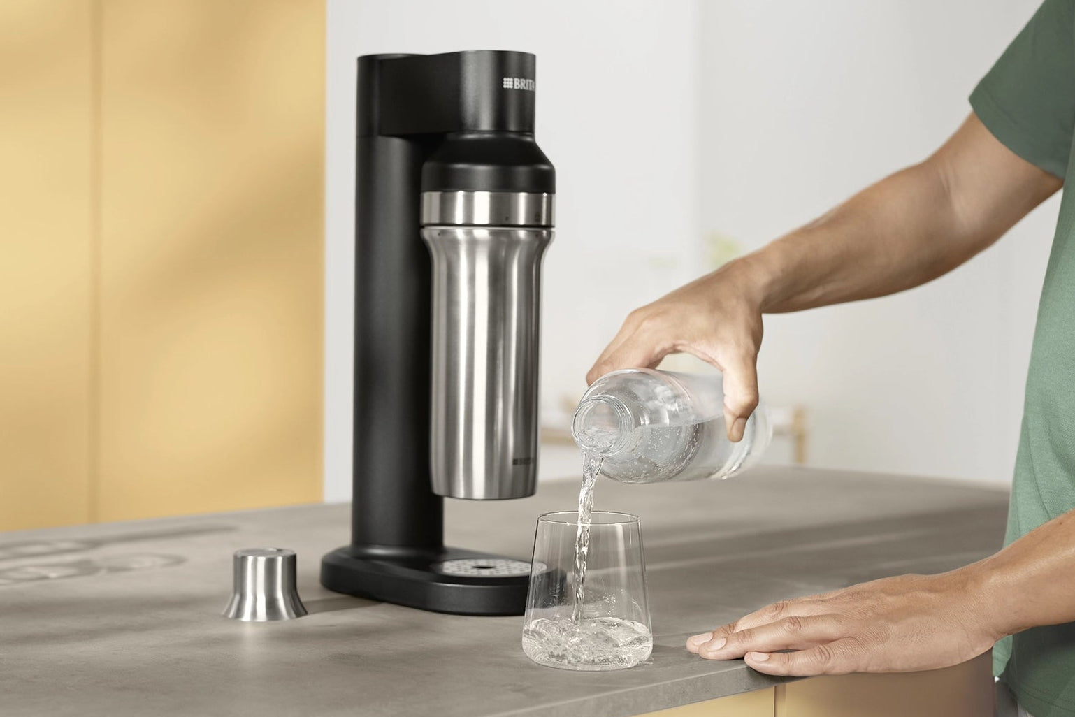 BRITA sodaTRIO Sparkling Water Maker with glass bottle pouring sparkling water into a glass on a kitchen counter.