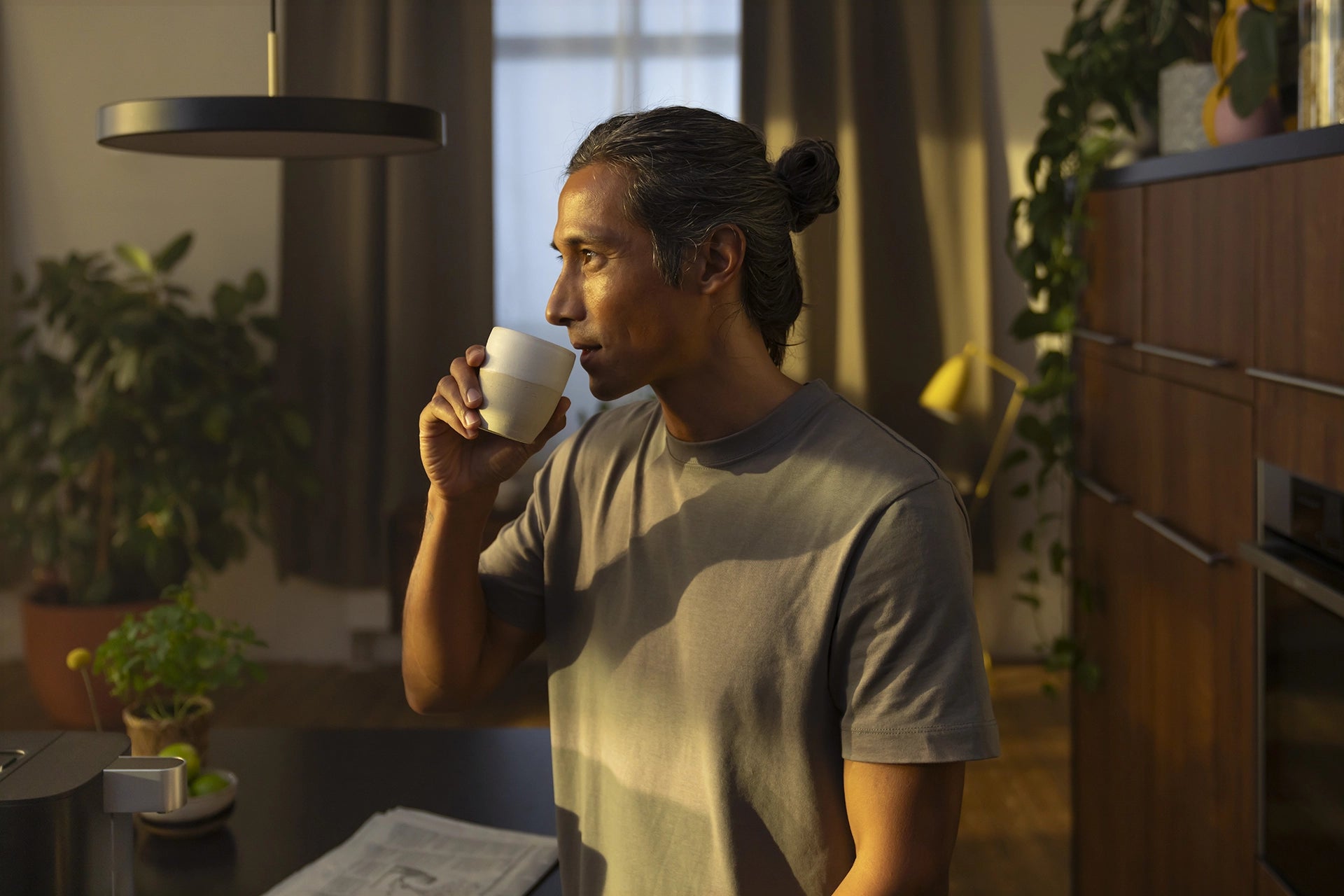Man drinking from a cup in a cozy kitchen with plants and furniture.