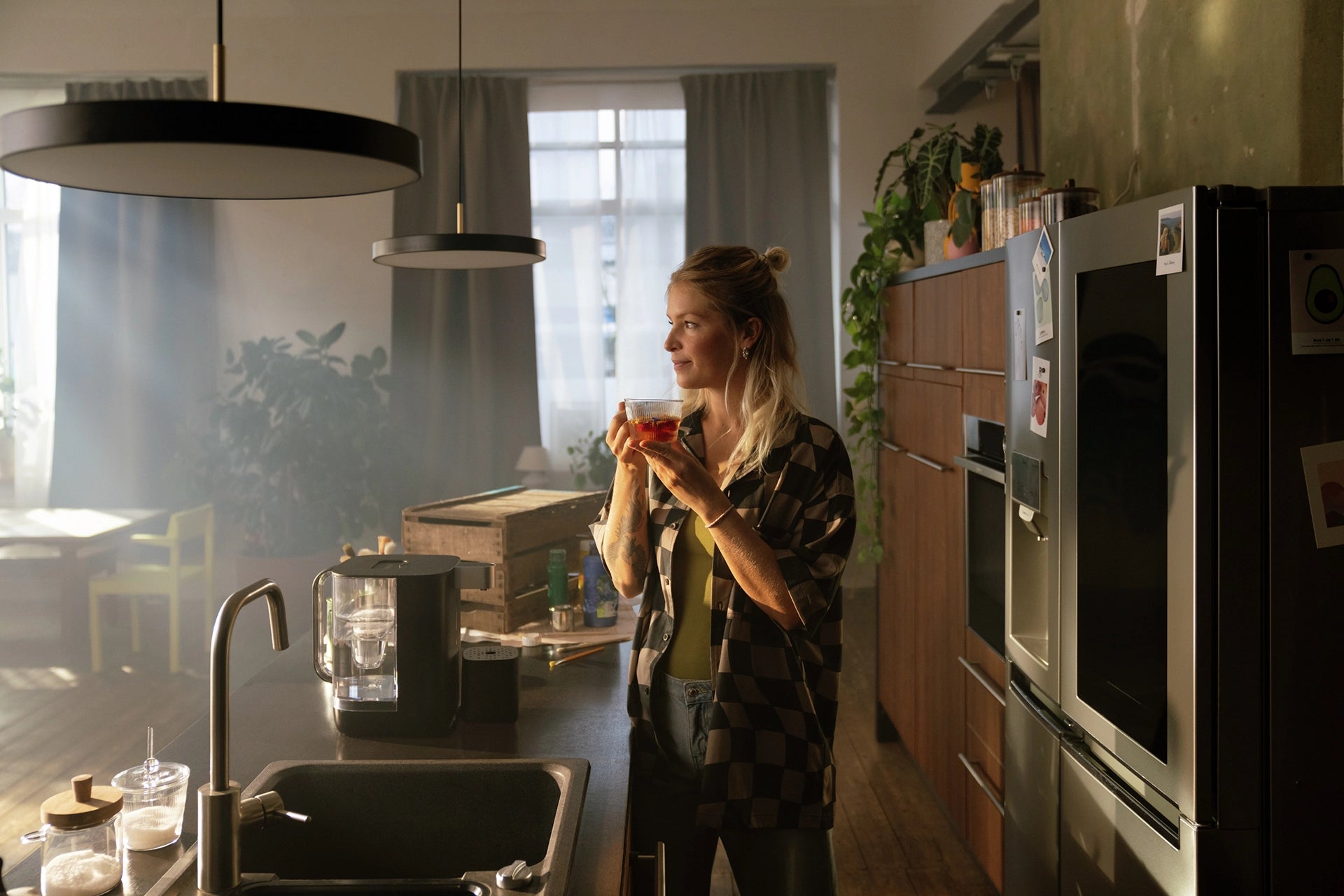 Person standing in a kitchen holding a glass cup of tea next to a BRITA YOURCE mini water dispenser.