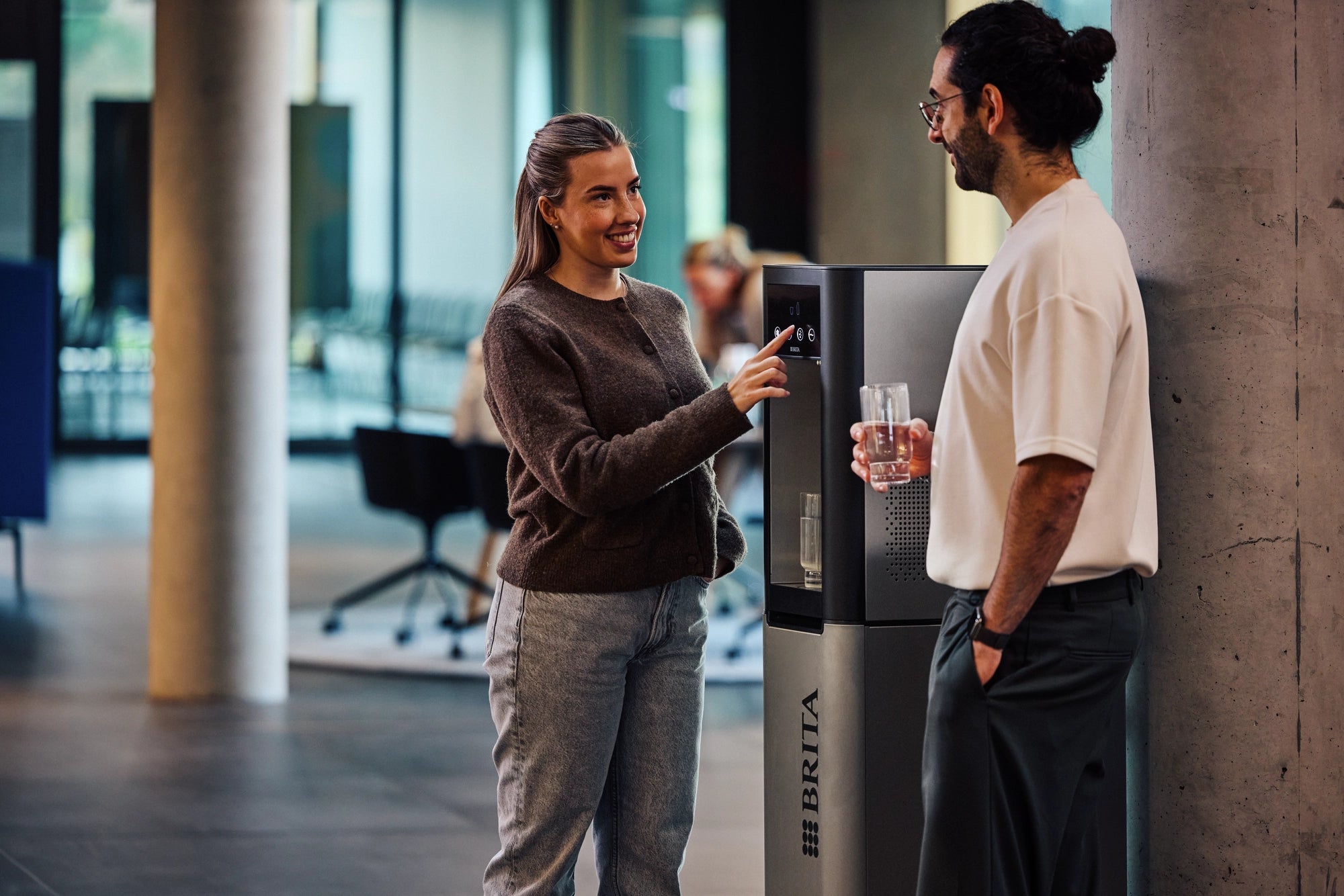 Two people using a BRITA waterdispenser while filling and holding glasses of water in an office environment.